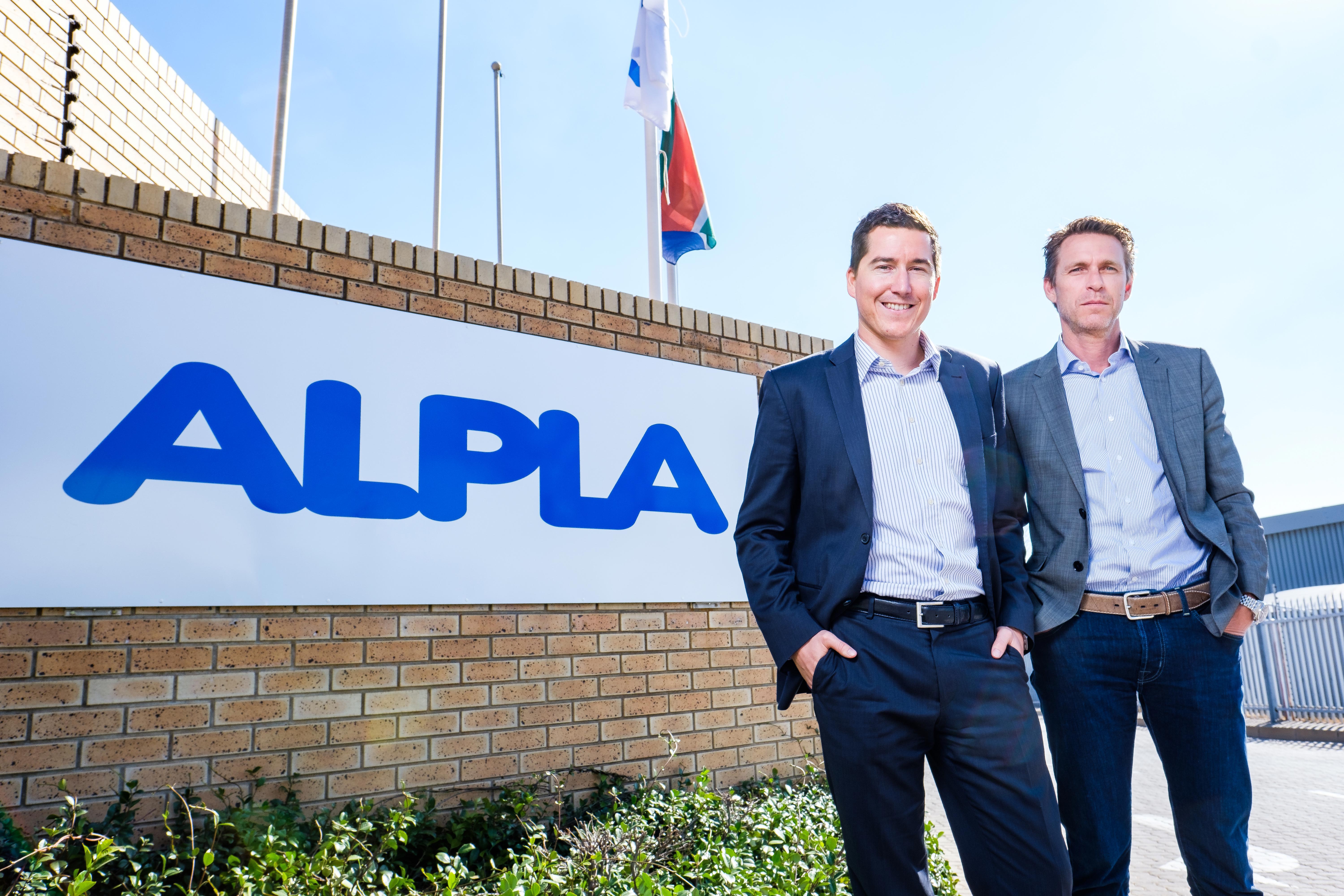 Two men in suits stand smiling by an ALPLA sign, under a clear sky.