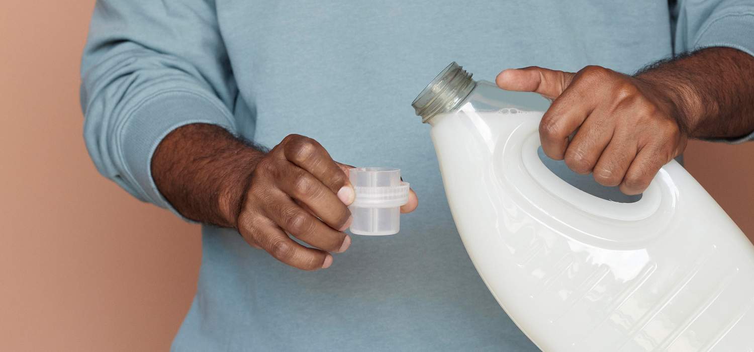 Person pouring liquid from white bottle into cap, wearing blue sweater.