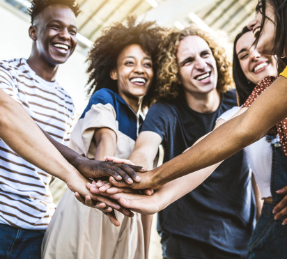 A group of young people standing in a circle, each placing their hand in the center of the group, high-fiving the back of the next person's hand. This energetic gesture creates a sense of unity and excitement, with everyone smiling or focused on the moment.