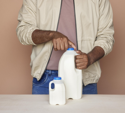 Mann mit zwei weißen Milchkannen, 500ml und 1 L, auf einem Tisch vor einem beigen Hintergrund. Tagebuchflaschen hergestellt von ALPLA