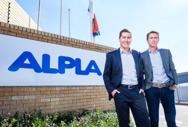 Two men in suits stand smiling by an ALPLA sign, under a clear sky.