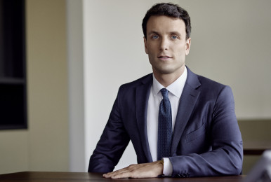Man in a suit seated at a desk in an office setting, looking composed.