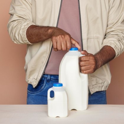 Man in beige jacket holding two white plastic milk jugs on a table. Manufactured by ALPLA.